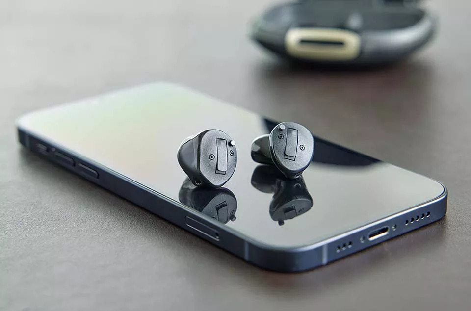 Two discreet in-ear hearing aids displayed beside a smartphone and charging case